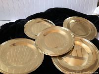 Five shiny gold tone charger plates arranged overlapping on a black background, showing reflective metallic finish and smooth, raised edged design.