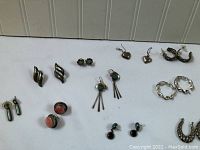 Overview of assorted earrings including small green drops, reddish stone studs, dangle earrings with rods, small turquoise studs, and twisted hoops.