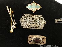 Five vintage brooches and pins displayed on black velvet background: golf clubs pin, filigree rectangle brooch, turquoise enamel quatrefoil brooch, garnet-colored stone oval brooch.