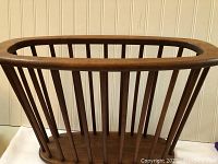 Front perspective view of an oval-top magazine holder with multiple vertical wooden slats and solid base against a paneled wall.