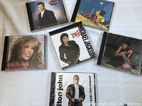 Six pop music CDs arranged on white surface showing cover art including Michael Jackson, Elton John, Carly Simon, Rick Astley, and two others with artwork.