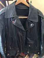 Front view of the black leather men's jacket hanging on a wooden hanger, showing zipper and snap button details on pockets and collar.