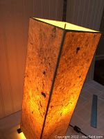 Lamp illuminated on a table, showing glowing textured natural fiber shade with square shape viewed from an angle.