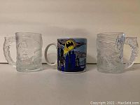 Three Batman coffee mugs displayed side by side on a flat surface with a plain background. Two clear glass mugs flank a ceramic mug with colored graphics.