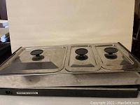 Electric buffet server with three rectangular stainless steel pans with lids, black knobs on lids, metal base with power switch, placed on a black surface against a plain background.