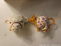 Shows two ceramic coffee mugs shaped like cows, one a white cow head with a nose ring and one a full-bodied cow wearing a yellow and floral patterned cloth with an orange handle.