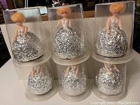 Six doll body cupcake picks displayed individually inside clear plastic containers, each doll has a foil skirt designed for cupcake insertion.