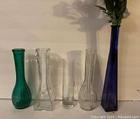 Photo showing five glass bud vases on a flat surface against a plain wall. The vases vary in height, shape, and color, with one tall cobalt blue vase, two clear glass vases, one teal vase, and one clear glass vase with a wider base and narrow opening.