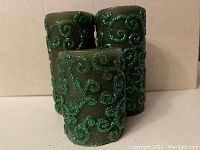 Three green pillar candles of different heights decorated with green glitter swirl patterns, arranged in a group on a neutral surface.