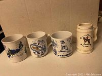 Four ceramic souvenir coffee mugs representing international locations: Nova Scotia, St. Kitts, Copenhagen, and Canada. Three standard mugs with destination artwork and one beer stein shape with moose antler handle depicting Canada.