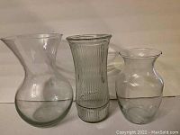 Photo showing three clear glass vases of different shapes and sizes on a flat surface against a plain background.
