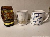 Photo showing three souvenir coffee mugs: one brown glass South Dakota Mt. Rushmore mug, one white ceramic Chicago skyline mug, and one white ceramic Chicago 'The Windy City' mug.