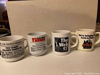 Four coffee mugs displayed in a single row in front of a cardboard background. Each mug features a different newspaper or radio station logo and advertisement from the 1990s.