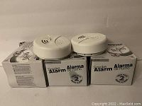 Photo showing six unopened white retail boxes labeled 'Smoke Alarm Alarma de humo' with two unpackaged white smoke alarms placed on top of the boxes. The alarms are round with a test button and vent openings.