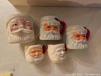 Photo showing five ceramic Santa Claus shaped coffee mugs with detailed facial features, white hats, red handles, and festive colors.