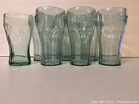 Photo of seven translucent green-tinted plastic Coca-Cola drinking cups arranged in two rows on a light surface with a beige background.