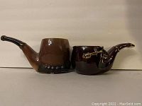 Photo showing two ceramic items shaped like smoking pipes used as ashtray or cups, both brown with glossy finishes, one larger with dark trim base and one smaller with metal pipe cleaner attachment.