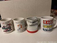 Photo of 4 white ceramic coffee mugs with humorous and snarky sayings printed on them, shown from the front in a row against a plain background.
