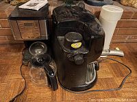 Photo showing three different coffee and tea makers on a wooden surface against a brick wall: a Mr. Coffee drip coffee maker, a black espresso machine, and a tall white electric kettle.