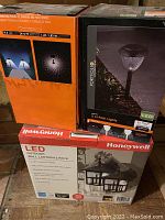 Three boxed sets of outdoor lighting stacked side by side showing product branding and images: Honeywell LED wall lanterns, Portfolio LED path lights, and a third LED light set with motion sensor.