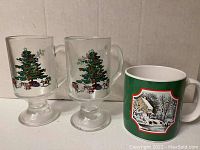 Photo of two clear glass mugs with Christmas tree and presents design, and one green ceramic coffee mug with a snowy house scene.