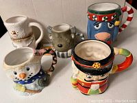 Photo showing five character-shaped winter/holiday coffee mugs including two snowmen, one nutcracker, one Eeyore, and one reindeer, arranged on a flat surface.
