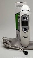 White digital forehead and ear thermometer with display and buttons, gray soft pouch, and original box in background.