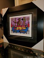Overall view of the professionally framed and matted artwork showing the colorful print and black frame with protective corner covers.