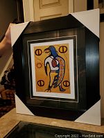 Framed and matted art print held upright showing overall presentation, including protective corner guards on frame.