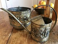 Photo showing side view of watering can with elongated spout and handle, brass with repousse rose and foliage design.