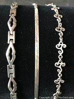 Photo shows all three bracelets side by side: snake link, flat swirl, and curly link design on black background.