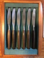 Set of six knives with stainless steel blades and dark teak wood handles inside a fitted green felt lined box.