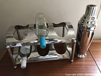 Wine rack with empty space for 6 bottles, with 2 wine bottles placed in rack, accompanied by cocktail shaker on right.