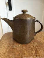 Front view of the teapot showing the spout, lid with knob, and handle against wooden surface and white background.