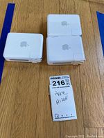 Three white Apple AirPort Base Stations arranged on a wooden surface with an identification tag showing model A1264.