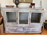 Front view of distressed wood and metal cabinet with three wire mesh doors and metal handles, distressed white finish, wooden top surface with natural grain.