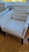 White leather upholstered chair with cylindrical lumbar pillow and low back cushion, showing front and top angles. Black wood legs, nailhead trim around edges.