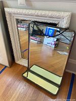 Two hanging mirrors, one with a metal frame and one with a white wood frame with visible damage, shown leaning against a wall on a wooden floor.