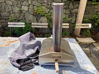 Front view of Uuni pizza oven on table with chimney and wooden handle visible. Grey Uuni cover and carrying strap beside it.