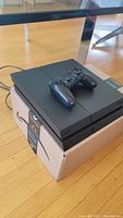 Sony PlayStation 4 console on top of a cardboard box, with a black DualShock 4 controller resting on it. Wooden floor and metal furniture legs visible.