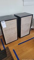 Two bookshelf speakers side by side showing beige fabric grilles and dark wood veneer sides.