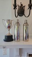 Full view of the vintage trophy and two stainless steel thermos bottles displayed on a white surface under a wall sconce.