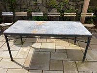 Front view of rectangular industrial outdoor metal table with heavily weathered metal top showing rust spots and black metal legs on a stone tiled patio.