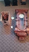 Handmade Gustav Becker German wall clock main body laid on carpet floor, showing front clock face and pendulum next to it. Cardboard box nearby holds detached parts.
