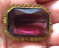 Hand-held image of the antique brooch showing the amethyst colored facetted glass with gilded brass filigree surrounding and old style back pin.