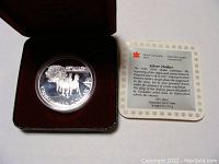 Silver coin inside black case with red velvet interior, showing detailed stagecoach design on one side with four horses and coachman.