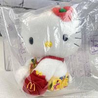 Front and side view of the Hello Kitty plush toy sealed in clear plastic bag showing red flower on ear, McDonald's apron, and floral skirt.