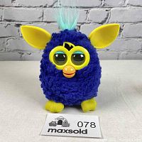 Front view of blue Furby with yellow feet and ears, teal hair tuft, large eyes, and orange beak displayed against white background.