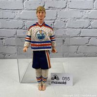 Front view of Wayne Gretzky doll in Edmonton Oilers white mesh jersey, blue shorts, and white socks with blue and orange stripes, standing barefoot in front of a brick wall.
