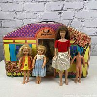 Four vintage dolls posed in front of the closed Tutti Playhouse case: one Barbie-sized with brown hair, two Tutti blonde dolls in outfits, and one undressed doll body without head.
